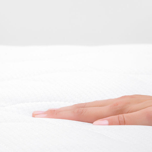 A woman pushing her hand into a mattress. She is testing how comfortable it is. There is a white wall in the background.