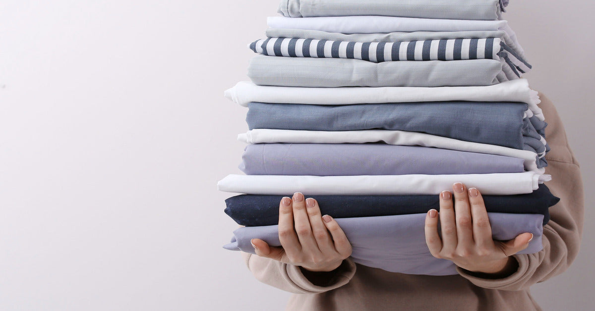 A woman holding a tall stack of bedsheets, covering her face. There are many different styles of sheets in the stack.