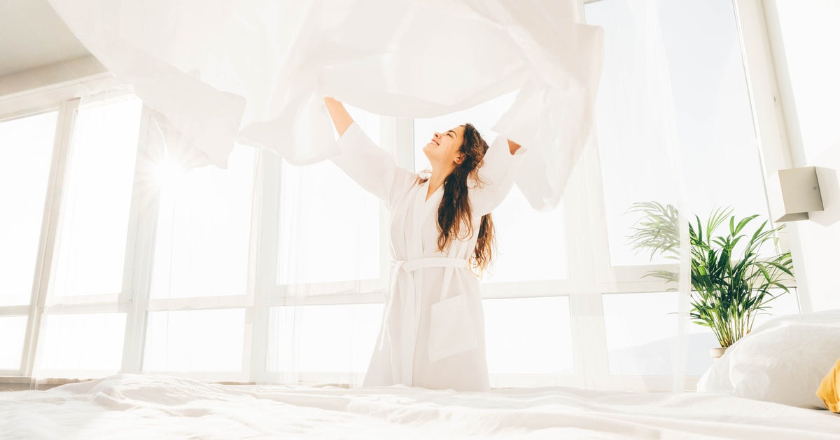 A woman throwing a white bed sheet in the air above her head. She's smiling while looking up at it.
