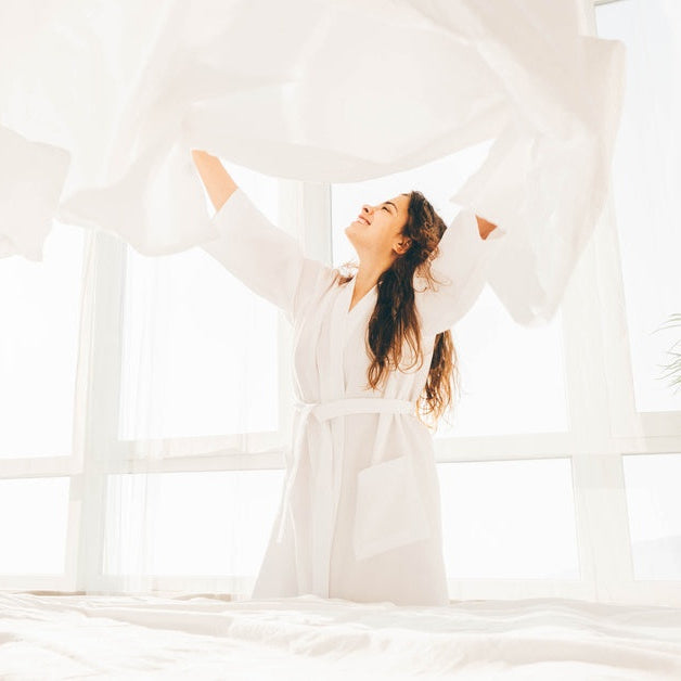 A woman throwing a white bed sheet in the air above her head. She's smiling while looking up at it.