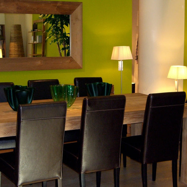 A plain wooden table with eight black chairs surrounding it. There is a mirror on the wall behind the table. 