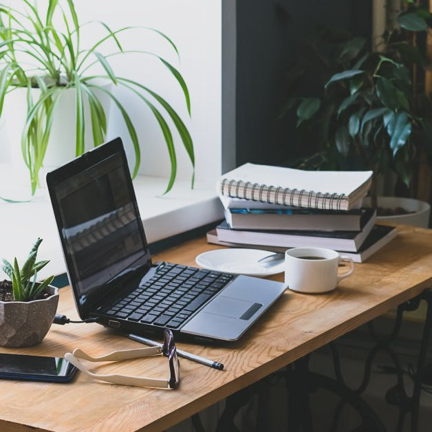 A wooden desk next to a window. There is a laptop, a stack of books, and other miscellaneous items on the desk.