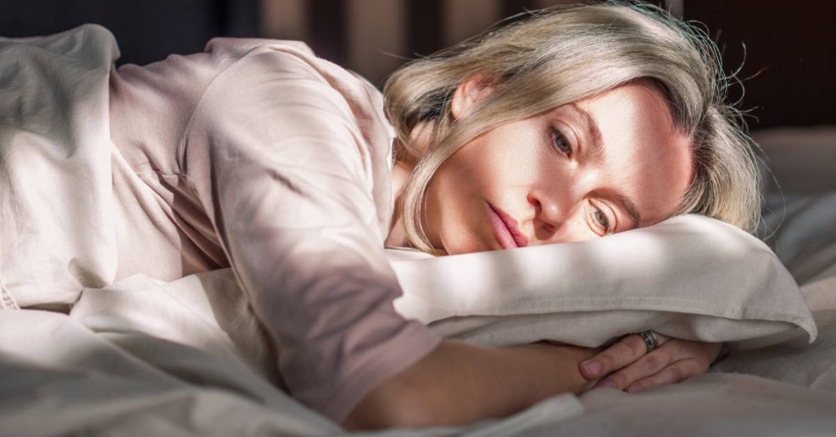 A woman laying on her bed with her arms wrapped around her pillow. The sun is shining through the binds and she looks unhappy.
