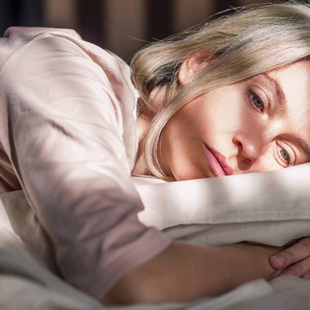 A woman laying on her bed with her arms wrapped around her pillow. The sun is shining through the binds and she looks unhappy.