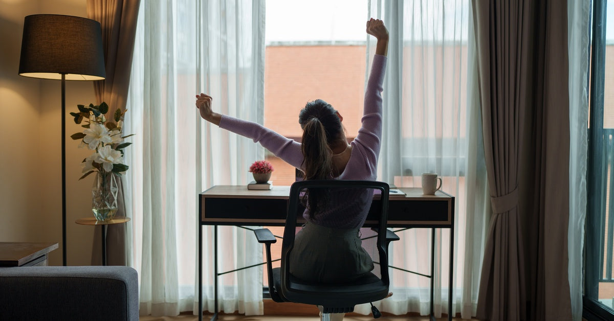 A woman sitting at her desk while stretching and looking out the window. She is using an ergonomic chair.