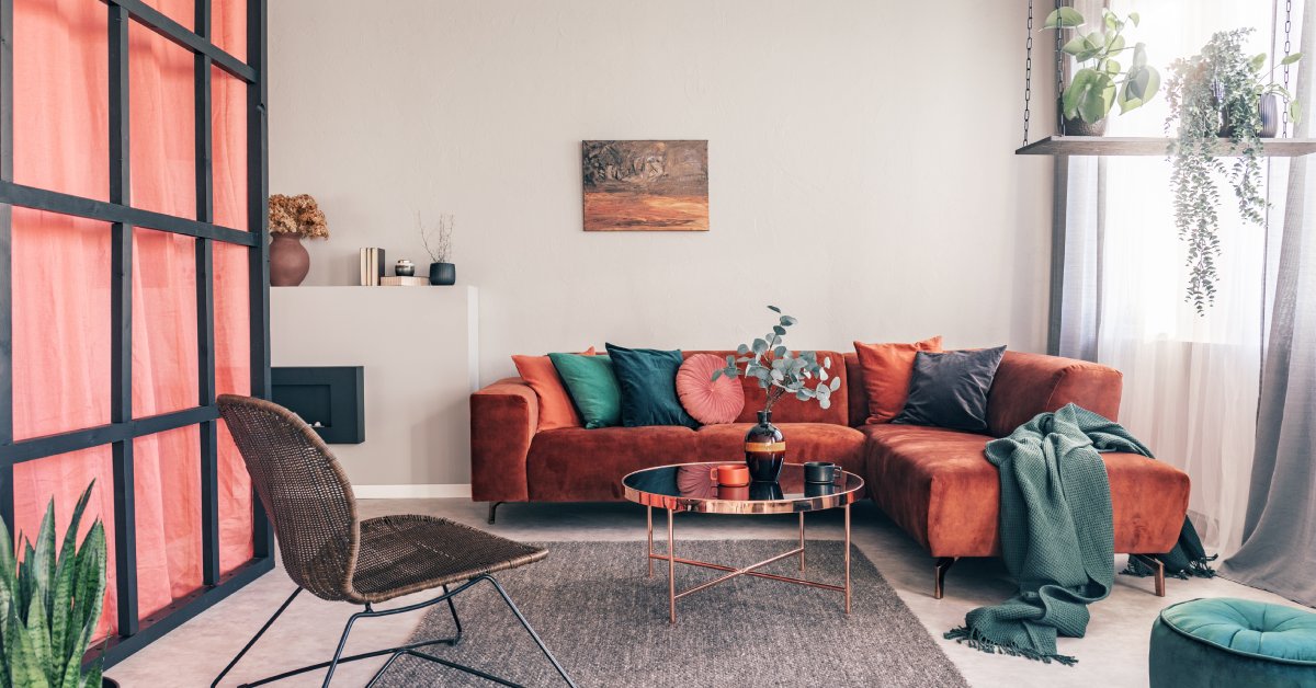 A living room with a red couch and wicker chair. The couch has a variety of pillows that are all different colors.