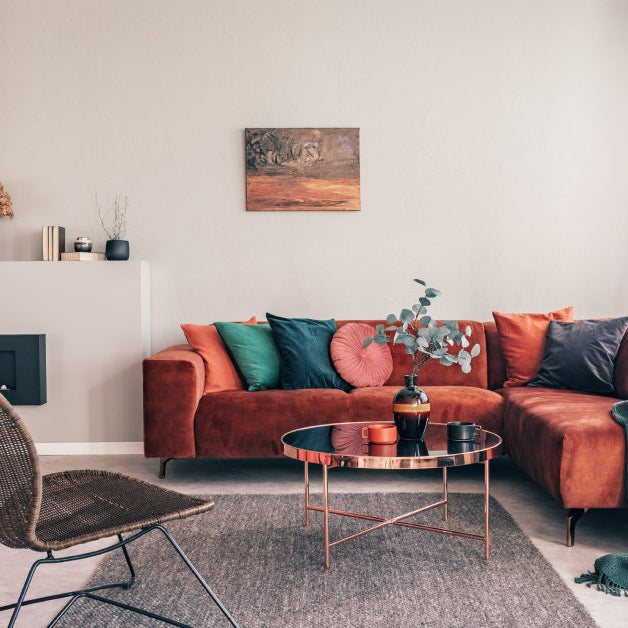A living room with a red couch and wicker chair. The couch has a variety of pillows that are all different colors.