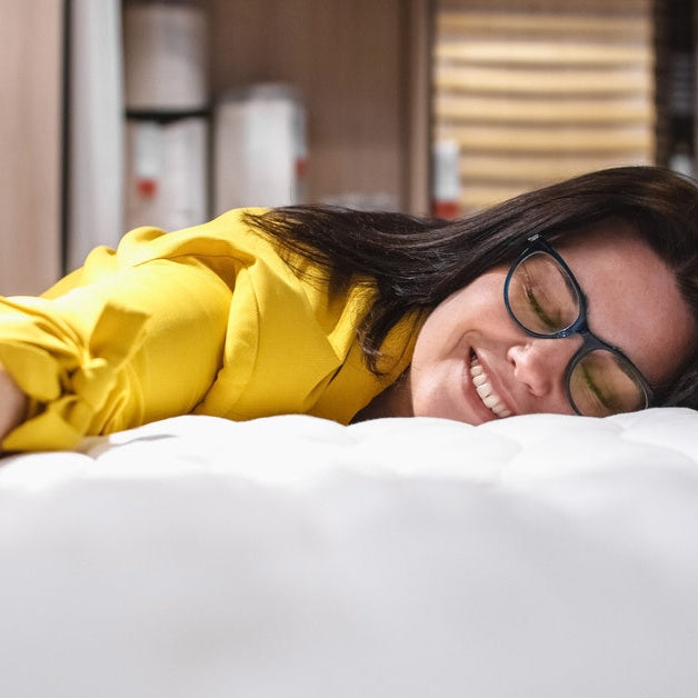A woman with glasses smiling as she lays down on her new mattress. She has her arms spread out across the surface.